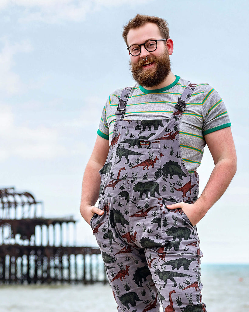 Thomas with a beard, glasses and nose ring is laughing stood by the West Pier in Brighton wearing grey dinosaur twill dungarees with a striped t-shirt underneath and black boots. He is facing the camera and is posing with both hands resting in the dungaree pockets.