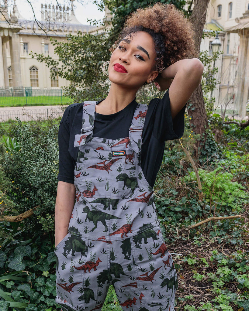 Cindy is wearing grey dinosaur run and fly dungaree overalls with turn up leg bottoms. She is wearing a short sleeved baggy black t-shirt underneath and shiny metallic black chunky boots. She is posing with her left hand in the pocket and her right hand behind her head, she is facing the camera and smiling.