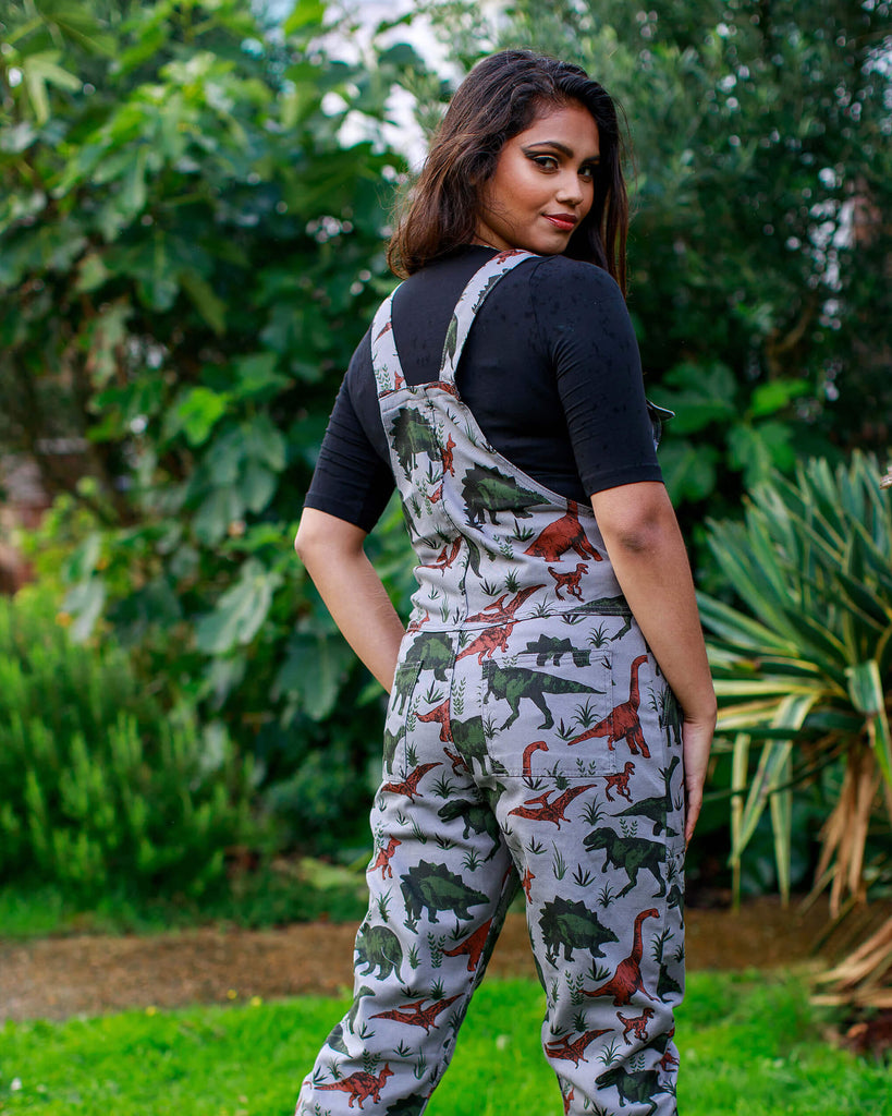 Jarin is wearing grey dinosaur run and fly dungaree overalls with turn up bottoms that were made in the UK. She is wearing a black mid length sleeve t-shirt underneath and black chunky boots. She is facing away from the camera and looking over her right shoulder into the lens.