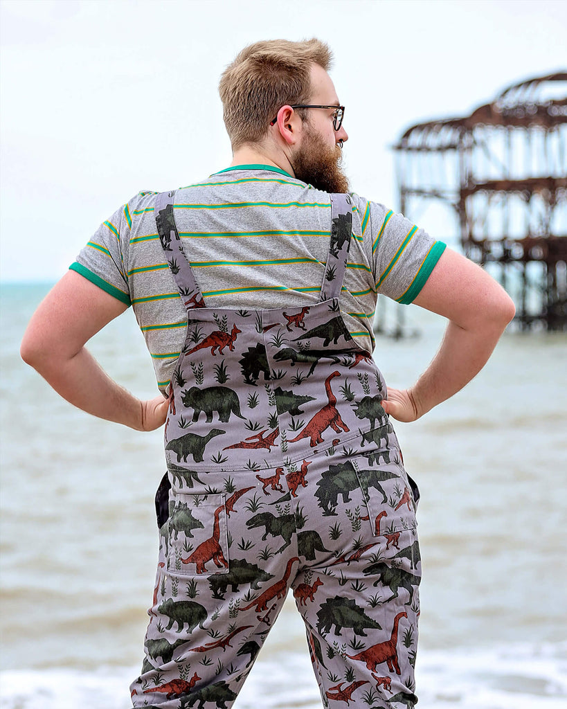 Man with a beard smiling by the West Pier in Brighton wearing grey dinosaur twill dungarees with a striped t-shirt underneath and black boots. He is facing away from the camera and posing looking to the right with both hands on his hips.