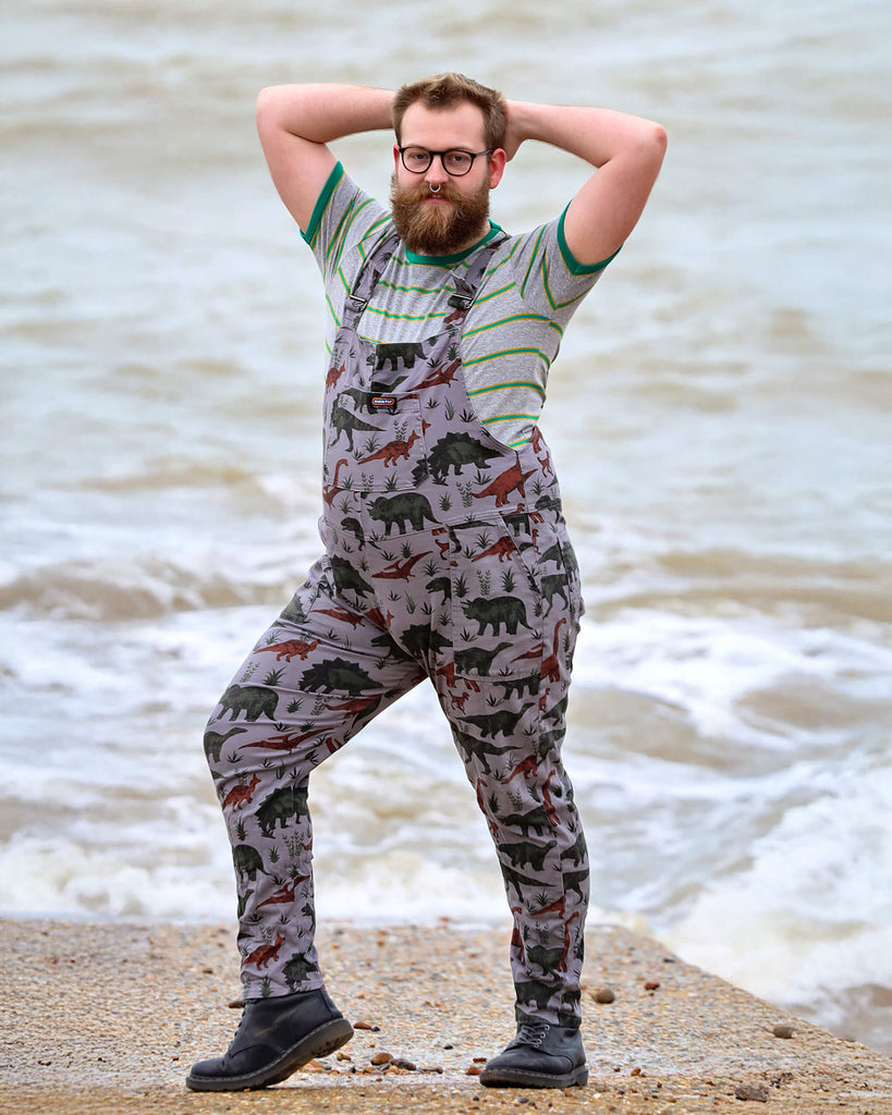 Thomas with a beard, glasses and nose ring is  smiling by the West Pier in Brighton wearing grey dinosaur twill dungarees with a striped t-shirt underneath and black boots. He is facing the camera and is posing with both hands behind his head and his left leg bent out the side.