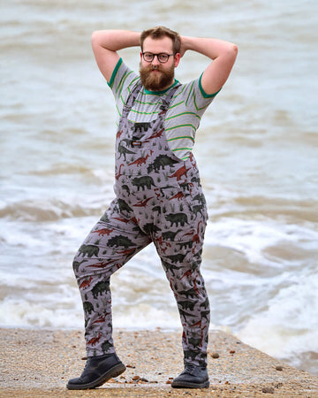 Thomas with a beard, glasses and nose ring is  smiling by the West Pier in Brighton wearing grey dinosaur twill dungarees with a striped t-shirt underneath and black boots. He is facing the camera and is posing with both hands behind his head and his left leg bent out the side.