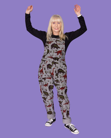 A woman with long blonde hair, dressed in grey corduroy dungarees with dinosaur prints and a black long-sleeve top, stands with her arms raised in excitement against a purple background.