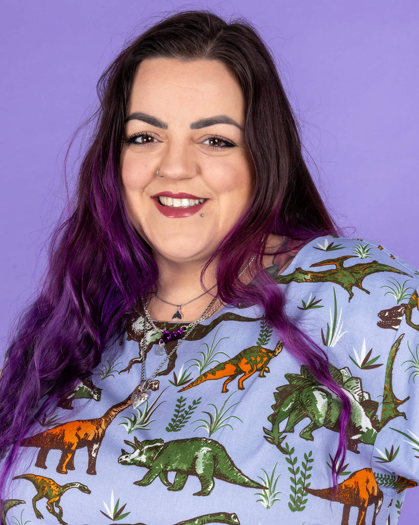 Luisa, a femme model with purple hair and tattoos, is stood in a photography studio in Hove in front of a purple backdrop wearing Jurassic Adventure Dinosaur Tea Dress With Pockets. The light blue dress has an all over dinosaur print with a matching fabric tie belt. Luisa is smiling at the camera and the photo is cropped at the waist.