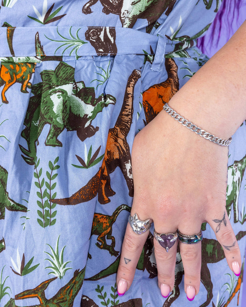 Close up of model's hands with tattoos and rings in the pocket of the Jurassic Adventure Dinosaur Tea Dress With Pockets.  The light blue dress has an all over dinosaur print with a matching fabric tie belt.