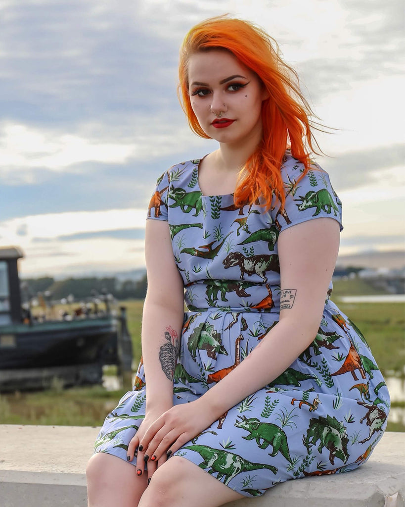 Tasha is sat on a brick wall wearing the jurassic adventure dinosaur tea party dress with black boots. She is looking into the camera and smiling with both hands in her lap.