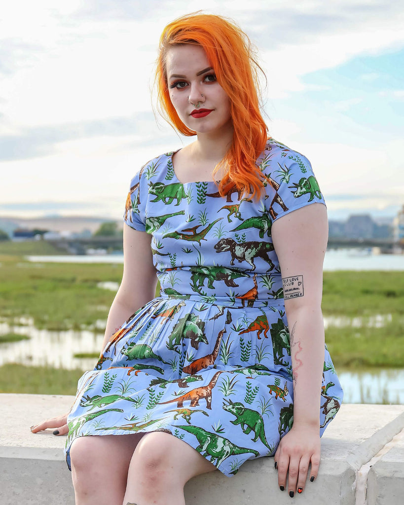 Tasha is sat on a brick wall wearing the jurassic adventure dinosaur tea party dress with black boots. She is looking into the camera and smiling with both hands resting on the wall she's sat on.