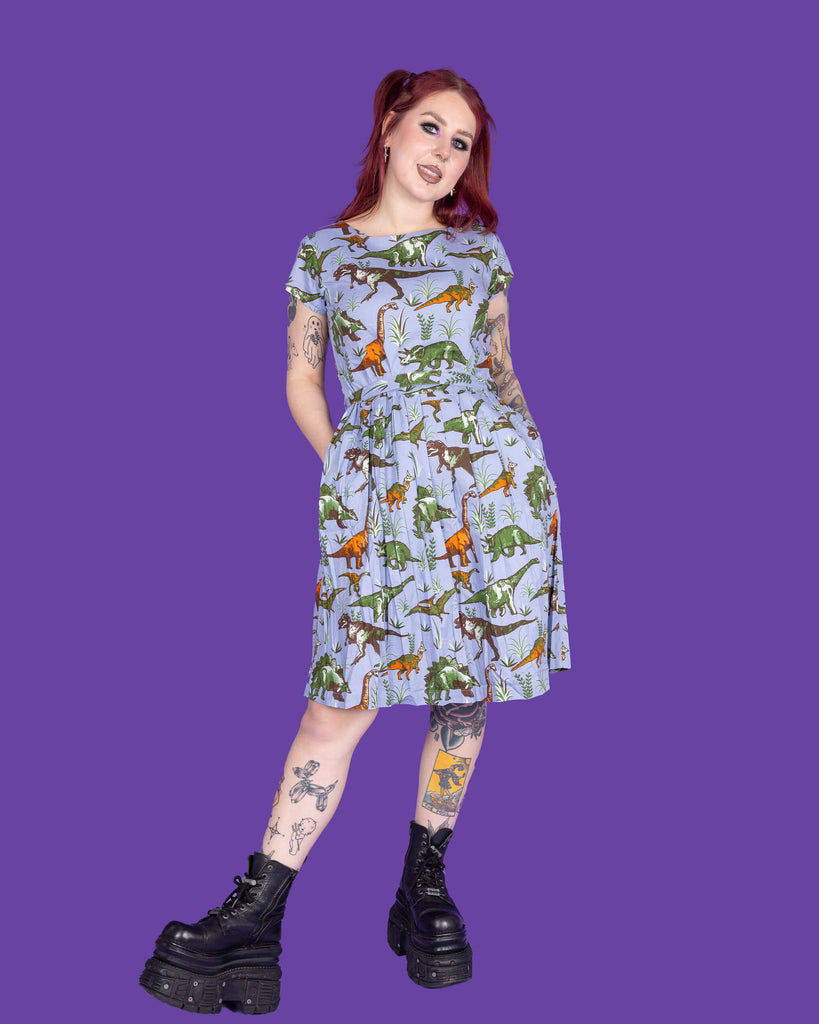 Florence, a tattooed model with red hair, is wearing Jurassic Adventure Dinosaur Tea Dress With Pockets paired with chunky black boots. The light blue dress has an all over dinosaur print with a matching fabric tie belt. Florence is stood in a studio in Hove in front of a purple backdrop posing toward the camera with both hands resting in the dress pockets.