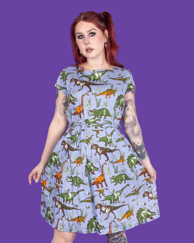 Florence, a tattooed model with red hair, is wearing Jurassic Adventure Dinosaur Tea Dress With Pockets paired with chunky black boots. The light blue dress has an all over dinosaur print with a matching fabric tie belt. Florence is stood in a studio in Hove in front of a purple backdrop posing toward the camera whilst holding the skirt of the dress.