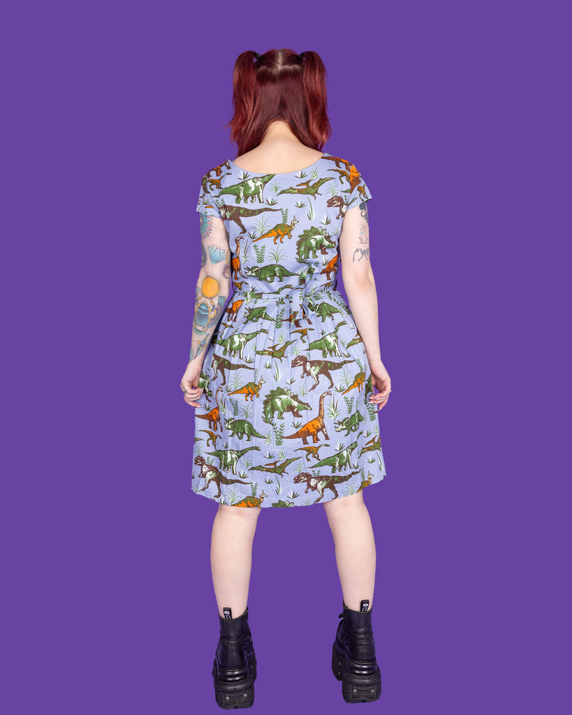 Florence, a tattooed model with red hair, is wearing Jurassic Adventure Dinosaur Tea Dress With Pockets paired with chunky black boots. The light blue dress has an all over dinosaur print with a matching fabric tie belt. Florence is stood in a studio in Hove in front of a purple backdrop posing with her back to the camera.