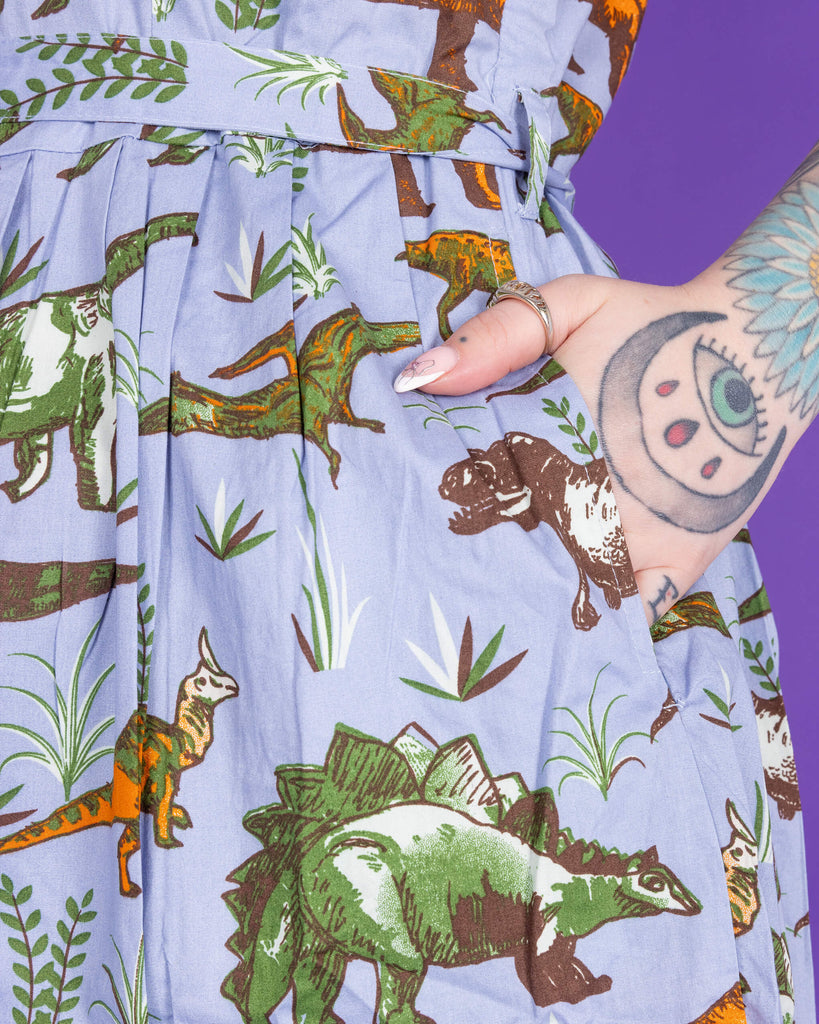 Close up of model's tattooed hand in pocket of Jurassic Adventure Dinosaur Tea Dress With Pockets. The background of the photo is purple.