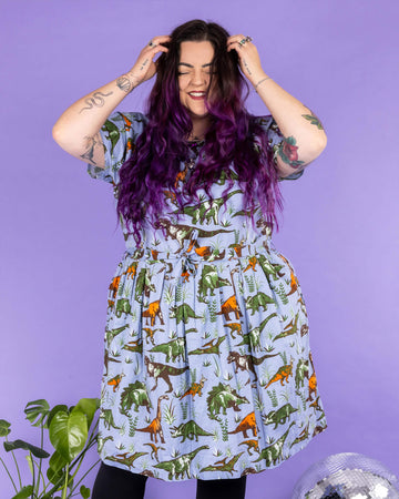 Luisa, a femme model with purple hair and tattoos, is stood in a photography studio in Hove in front of a purple backdrop wearing Jurassic Adventure Dinosaur Tea Dress With Pockets with black tights and boots. The light blue dress has an all over dinosaur print with a matching fabric tie belt. Luisa is stood on a black furry rug amongst disco balls, a star light and a plant and is facing the camera smiling with her hands in her hair.