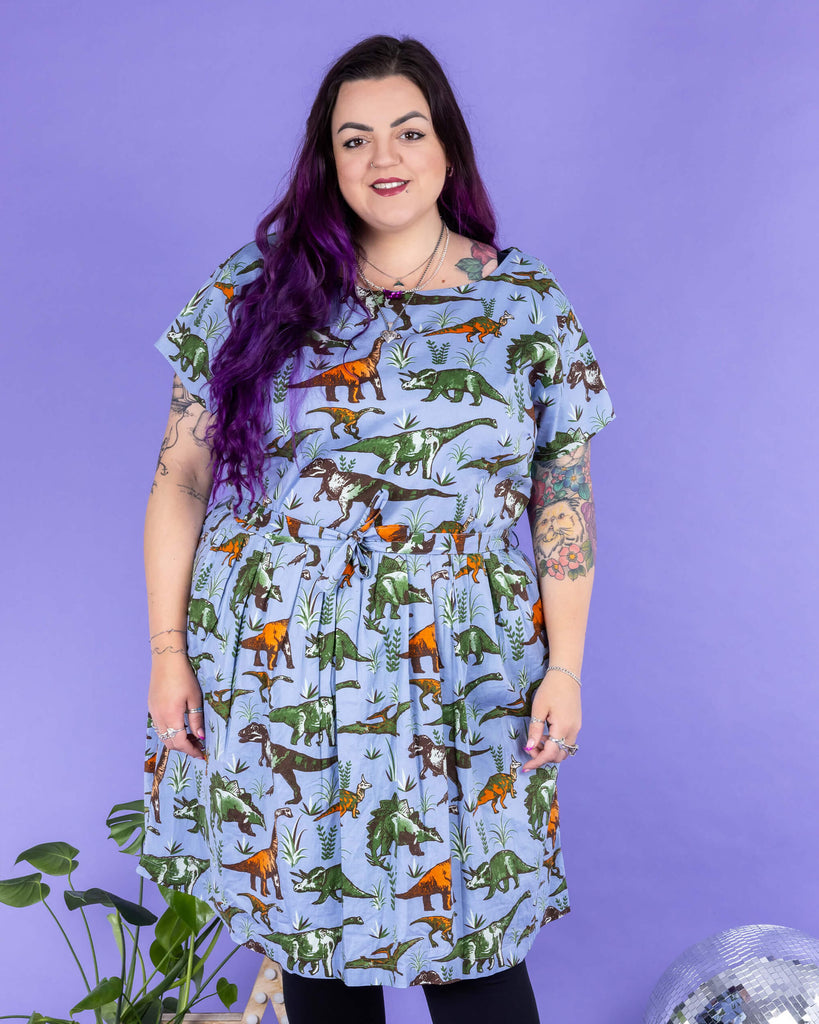 Luisa, a femme model with purple hair and tattoos, is stood in a photography studio in Hove in front of a purple backdrop wearing Jurassic Adventure Dinosaur Tea Dress With Pockets with black tights and boots. The light blue dress has an all over dinosaur print with a matching fabric tie belt. Luisa is stood on a black furry rug amongst disco balls, a star light and a plant and is posing toward the camera with her arms down by her sides and one leg in front of the other.