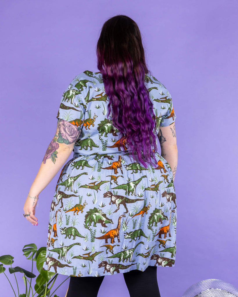 Luisa, a femme model with purple hair and tattoos, is stood in a photography studio in Hove in front of a purple backdrop wearing Jurassic Adventure Dinosaur Tea Dress With Pockets with black tights and boots. The light blue dress has an all over dinosaur print with a matching fabric tie belt. Luisa is stood on a black furry rug amongst disco balls, a star light and a plant with her back to the camera.