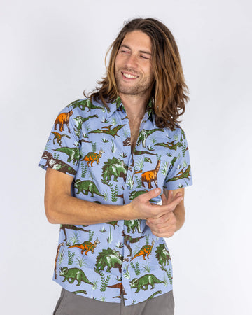 Model wearing a short-sleeved dinosaur-themed shirt from Run & Fly, a small ethical slow fashion company. The light blue shirt is adorned with vibrant green, orange, and brown dinosaurs. The model is smiling and looking off to the side, showing off the fun print and comfortable, casual fit. Ideal for those who love unique, retro-inspired style with an adventurous twist.