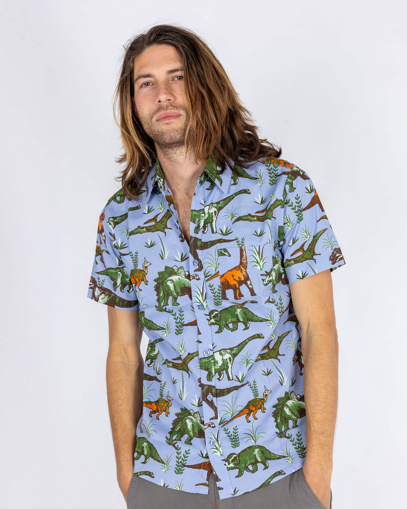 Front-facing shot of a model wearing a light blue short-sleeved dinosaur shirt by Run & Fly, a small ethical slow fashion company. The shirt’s print features green, orange, and brown dinosaurs set against a light blue backdrop. The model’s relaxed stance showcases the shirt’s easy, laid-back fit, ideal for vintage lovers and dinosaur enthusiasts alike.
