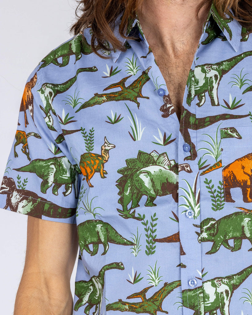 Close-up of the Run & Fly dinosaur print shirt, highlighting the intricate design. The light blue shirt features an array of green, orange, and brown dinosaurs, with plants and trees, in a fun, detailed print. Made by a small ethical slow fashion company, this playful shirt adds a retro flair to any wardrobe. Perfect for those seeking bold, quirky styles.