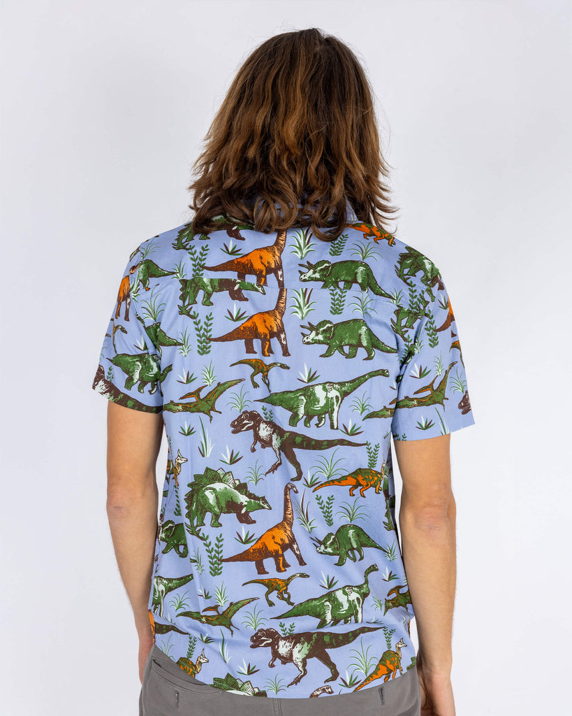 Rear view of the Run & Fly light blue dinosaur print shirt, showcasing the playful design in full. The print features green, orange, and brown dinosaurs against a light blue background. The short-sleeved shirt has a relaxed fit, perfect for casual wear. Made by a small ethical slow fashion company, it’s ideal for anyone who loves fun, quirky fashion with a retro vibe.