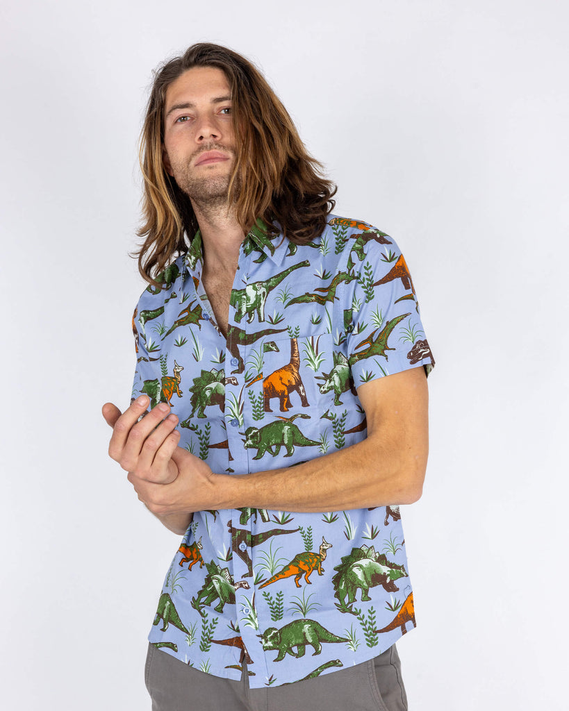 Model in a short-sleeved light blue button-up shirt featuring a playful dinosaur print in green, orange, and brown, standing with arms loosely crossed. The shirt is by Run & Fly, a small ethical slow fashion company, known for bold, quirky patterns. The model’s casual pose highlights the shirt’s relaxed fit, perfect for making a statement with a vintage-inspired look.