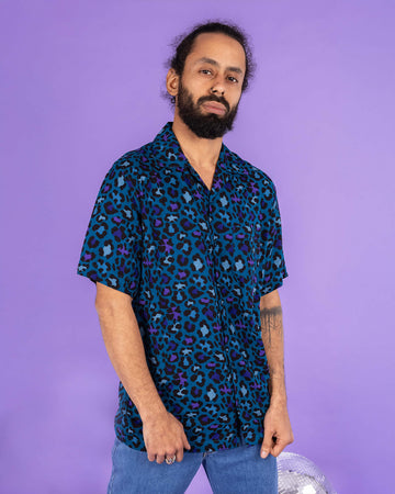 Richard, a hispanic male model with dark hair in a bun and a beard, is stood in a photography studio in Hove in front of a lilac backdrop wearing Atlantic Blue Leopard Print Short Sleeve Rayon Shirt with blue jeans. Richard is posing to the camera with his hands resting at the bottom of the shirt. The photo is cropped at the thighs.