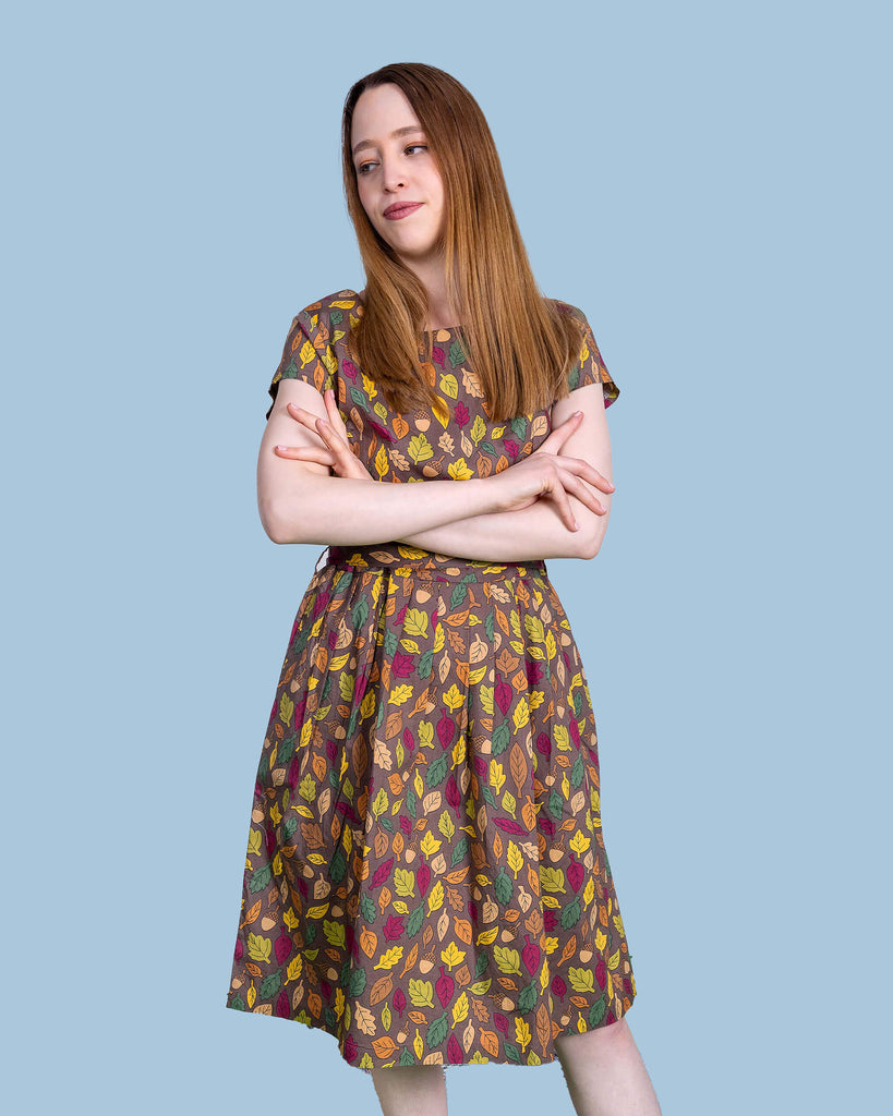 The Autumn Leaves Stretch Belted Tea Dress with Pockets on a femme model with long brown hair and black heeled boots. She is posing leaning back on one leg with her arms crossed smiling to the left at a picnic setup with a picnic basket, fruit, flowers and katie abey mugs. The dress print is a brown base with green, red, orange and yellow autumnal leaves and acorns.