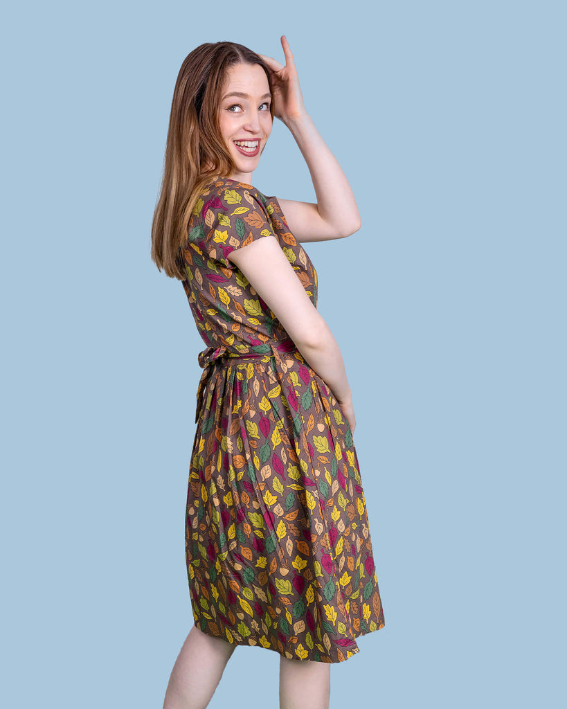 The Autumn Leaves Stretch Belted Tea Dress with Pockets on a femme model with long brown hair and black heeled boots. She is posing facing away laughing looking over her shoulder up to the left with her hand resting on her head at a picnic setup with a picnic basket, fruit, flowers and katie abey mugs. The dress print is a brown base with green, red, orange and yellow autumnal leaves and acorns.