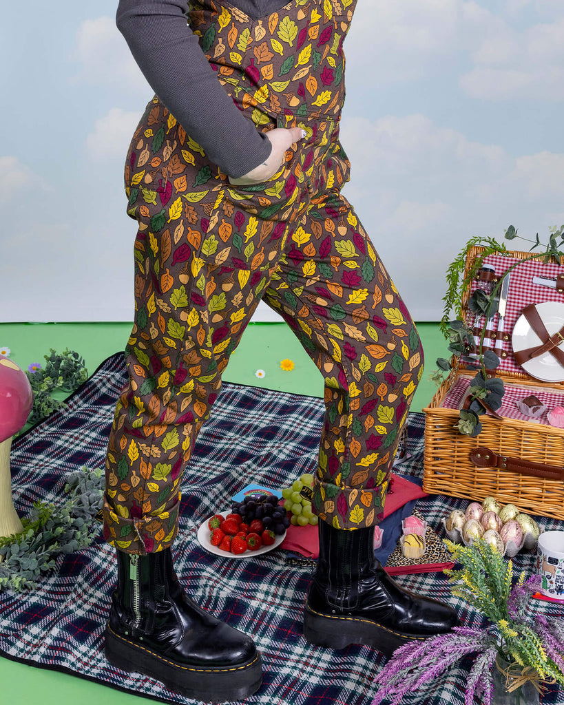 Close up of Claudia's legs standing facing right with both hands in the dungaree pockets wearing brown based dungarees covered in leaves and acorn illustrations. She is stood in front of a picnic scene.