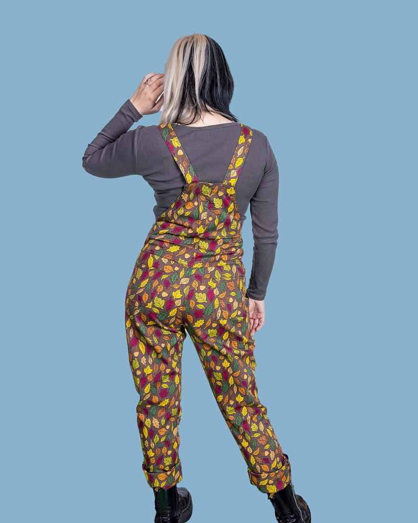 Claudia is a white model with half black and half blonde hair. She is looking away from camera wearing brown based dungarees covered in leaves and acorn illustrations. She is stood in front of a blue background.