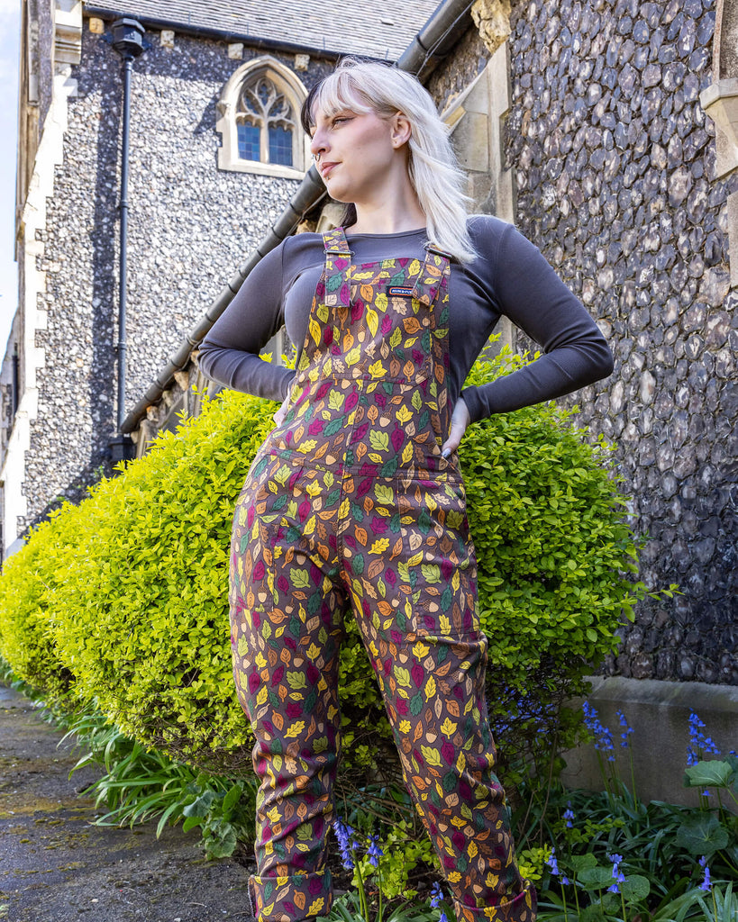 Claudia is a white model with half black and half blonde hair. She is looking into camera and smiling wearing brown based dungarees covered in leaves and acorn illustrations. She is stood in front of a church outside 