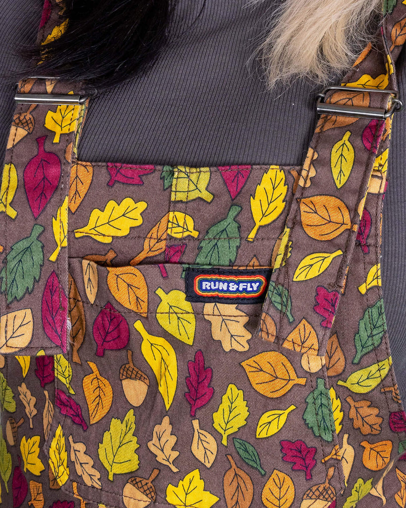 close up of the bib of Claudia a white model with half black and half blonde hair wearing brown based dungarees covered in leaves and acorn illustrations. 