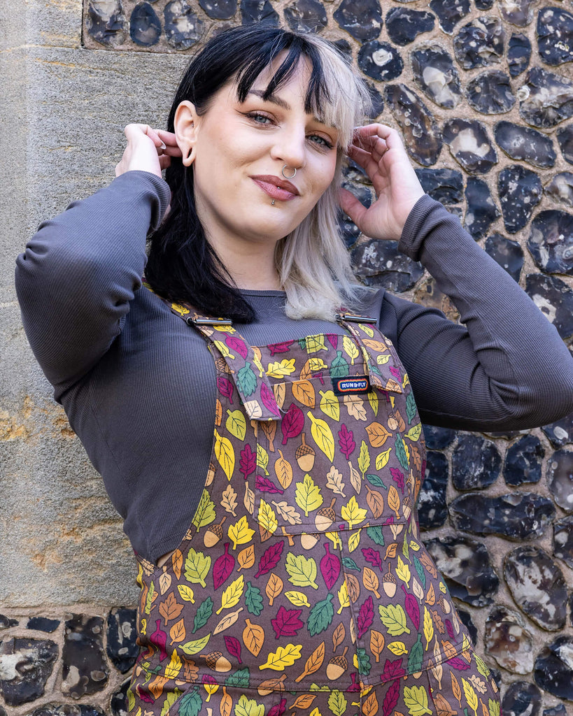 Claudia is a white model with half black and half blonde hair. She is looking into camera and smiling wearing brown based dungarees covered in leaves and acorn illustrations. She is stood in front of a church outside 