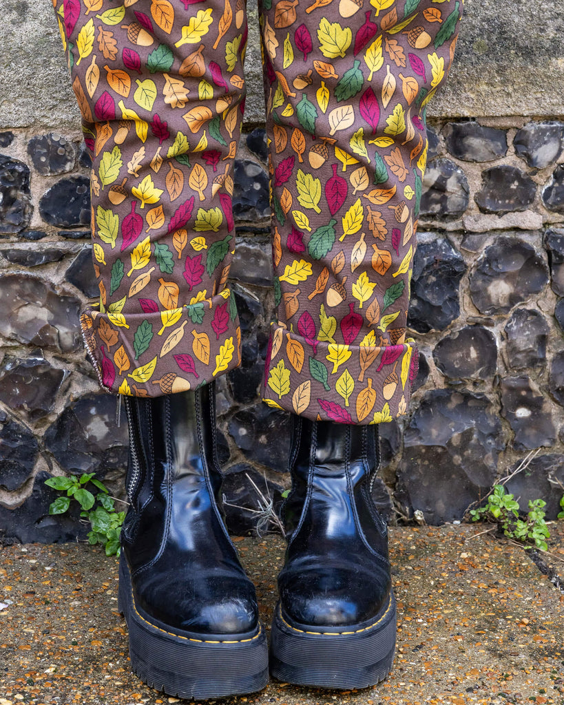 Close up of the turn ups of autumn leaves dungarees paired with black boots on Claudia stood outside in front of a brick wall. Brown based dungarees covered in leaves and acorn illustrations.