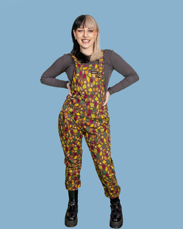Claudia is a white model with half black and half blonde hair. She is looking into camera and smiling wearing brown based dungarees covered in leaves and acorn illustrations. She is stood on a light blue background.