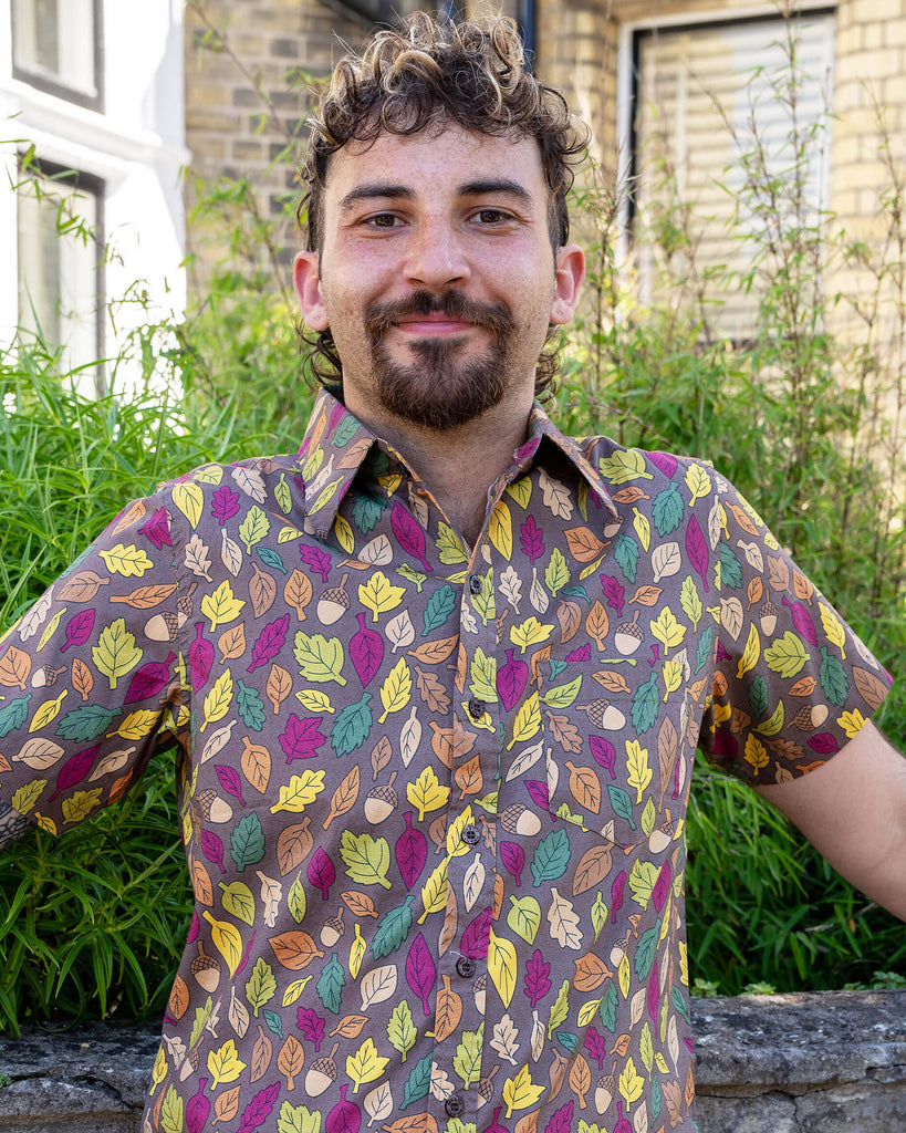 The Autumn Leaves Short Sleeve Shirt worn by a masc non binary person with a curly brown mullet cut and denim shorts. They are stood outside along the pavement in front of a bush and ornate wall facing forward smiling leaning back on the wall. The light brown based shirt features dark brown, yellow, orange, dark red, dark green and beige leaves with acorns all over. 