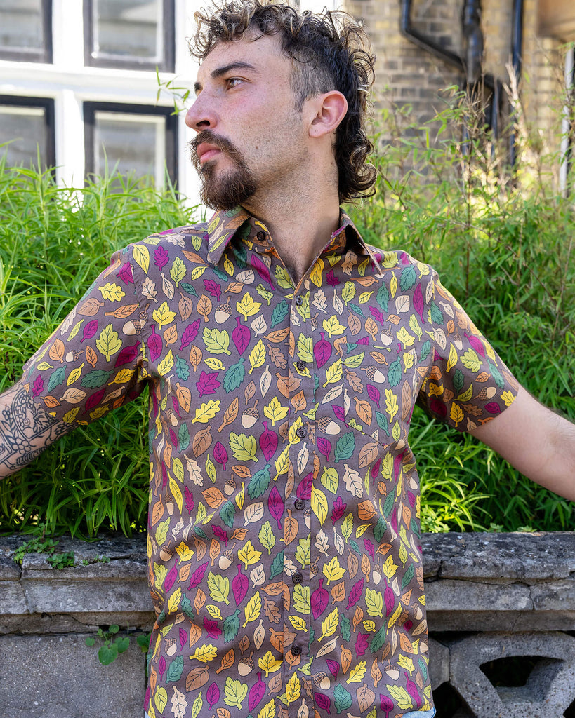 The Autumn Leaves Short Sleeve Shirt worn by a masc non binary person with a curly brown mullet cut and denim shorts. They are stood outside along the pavement in front of a bush and ornate wall facing forward looking off to the left leaning back on the wall. The light brown based shirt features dark brown, yellow, orange, dark red, dark green and beige leaves with acorns all over.