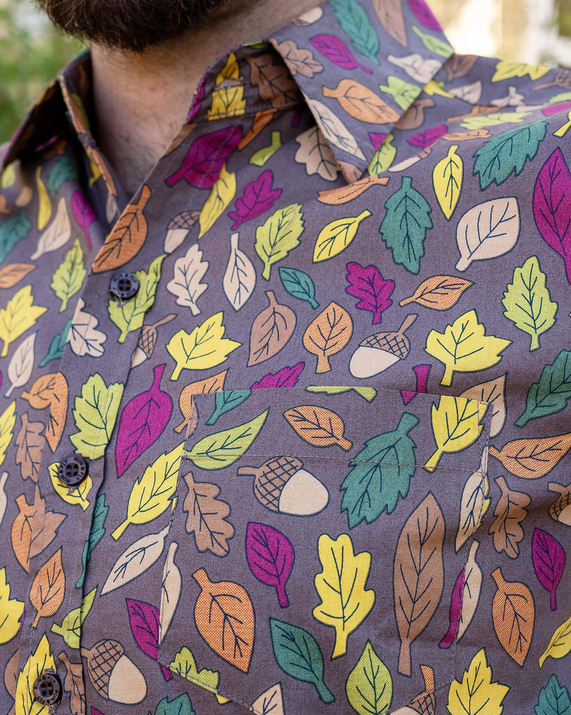 Close up of the Autumn Leaves Short Sleeve Shirt worn by a masc non binary person. The light brown based shirt features dark brown, yellow, orange, dark red, dark green and beige leaves with acorns all over.