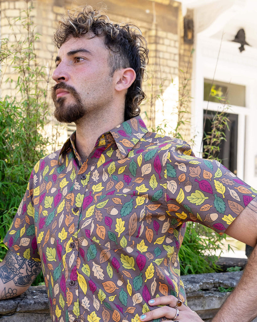 The Autumn Leaves Short Sleeve Shirt worn by a masc non binary person with a curly brown mullet cut and denim shorts. They are stood outside along the pavement in front of a bush and ornate wall facing forward leaning on the wall with one hand on their hip looking to the left. The light brown based shirt features dark brown, yellow, orange, dark red, dark green and beige leaves with acorns all over.