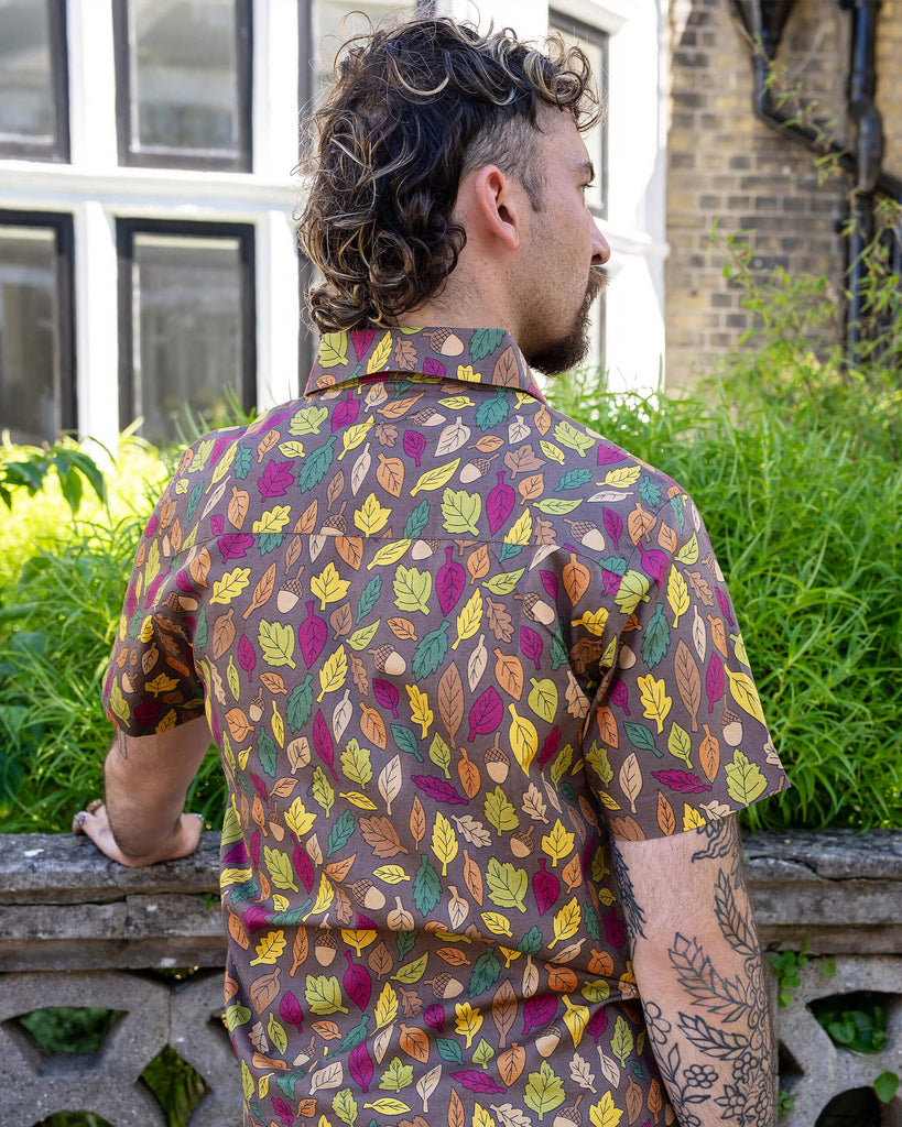 The Autumn Leaves Short Sleeve Shirt worn by a masc non binary person with a curly brown mullet cut and denim shorts. They are stood outside along the pavement in front of a bush and ornate wall facing away with one hand resting on the wall and looking slightly over their shoulder to the right. The light brown based shirt features dark brown, yellow, orange, dark red, dark green and beige leaves with acorns all over.