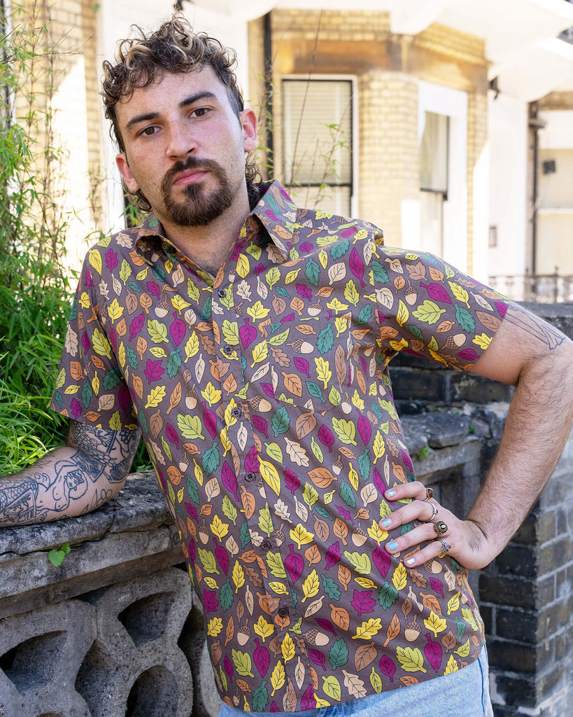 The Autumn Leaves Short Sleeve Shirt worn by a masc non binary person with a curly brown mullet cut and denim shorts. They are stood outside along the pavement in front of a bush and ornate wall facing forward leaning on the wall with one hand on their hip looking to camera. The light brown based shirt features dark brown, yellow, orange, dark red, dark green and beige leaves with acorns all over.