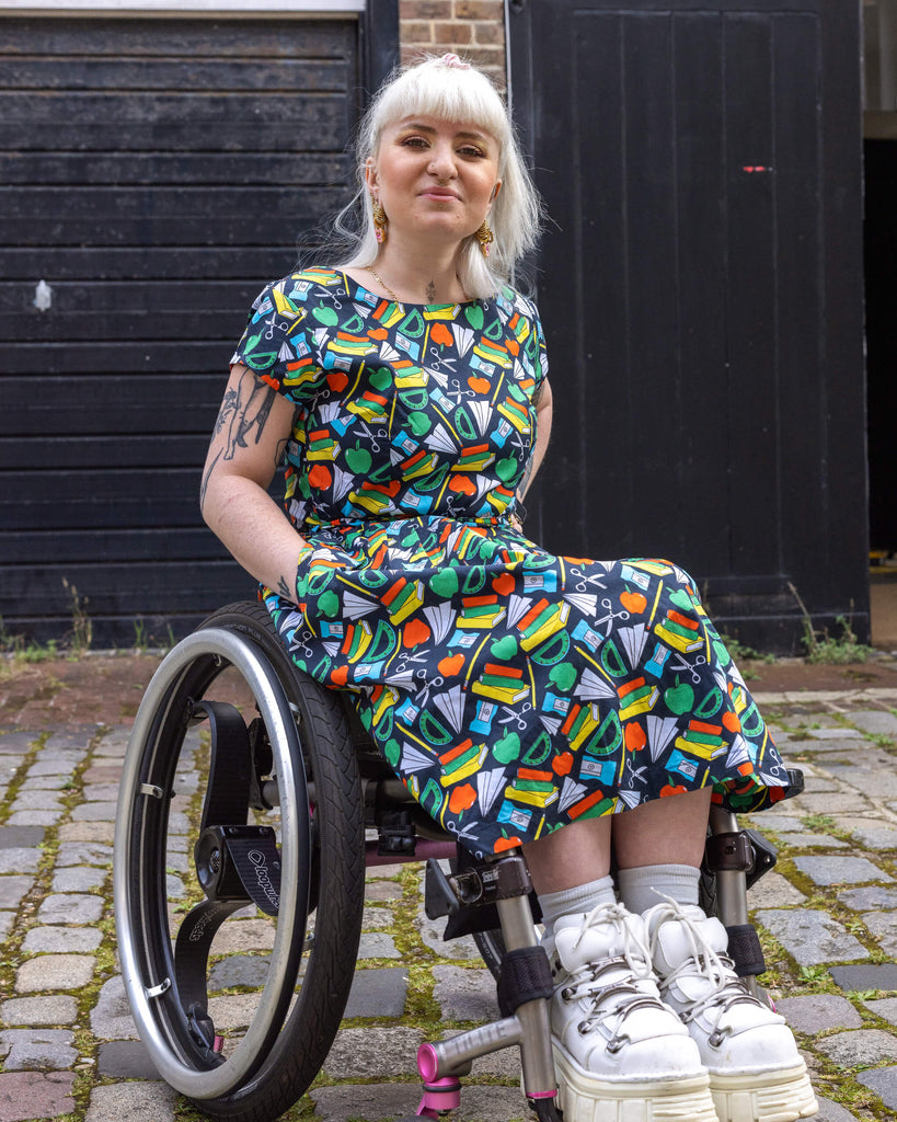 Model posing  in a wheelchair wearing Back to School Stretch Belted Tea Dress with Pockets on a cobblestone path. The dress is detailed with a vibrant array of educational motifs like books, rulers, paper planes, and apples, all in bright colours against a navy blue background.