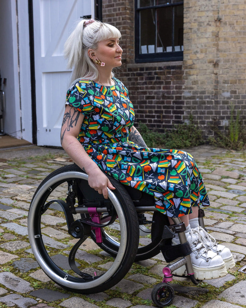 Side profile of a model wearing Back to School Stretch Belted Tea Dress with Pockets in a wheelchair. The pattern includes books, rulers, apples, paper planes, and scissors in vibrant colours on a dark navy background. 