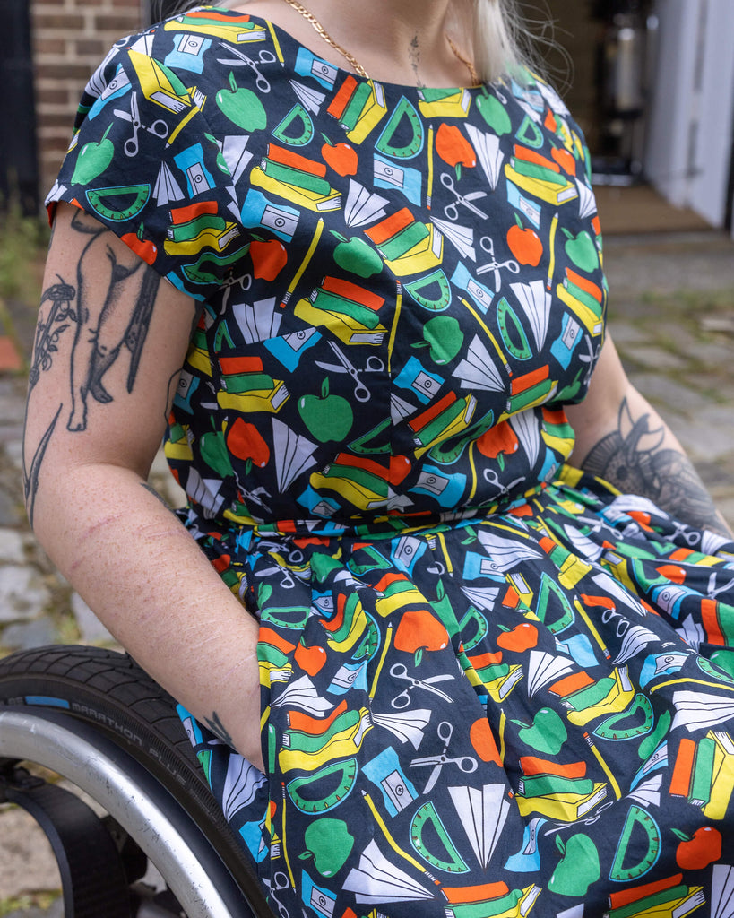 Detailed shot of Back to School Stretch Belted Tea Dress with Pockets worn by a model in a wheelchair. The dress is adorned with colourful illustrations of books, rulers, apples, and scissors on a navy blue backdrop. The close up shows the models hand in the pocket of the dress