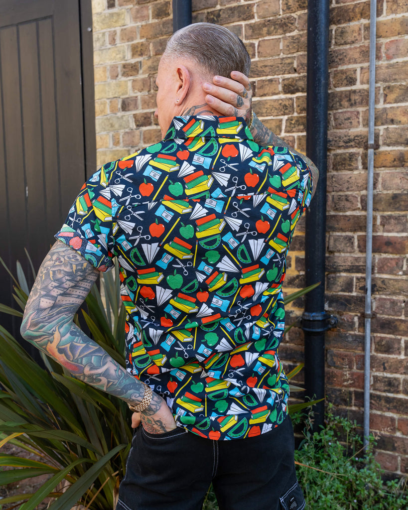 An older man with tattoos models a vibrant short-sleeve shirt featuring a pattern of school supplies on a dark base colour. He stands against a brick wall, smiling and looking to the side with his hands on his hips.