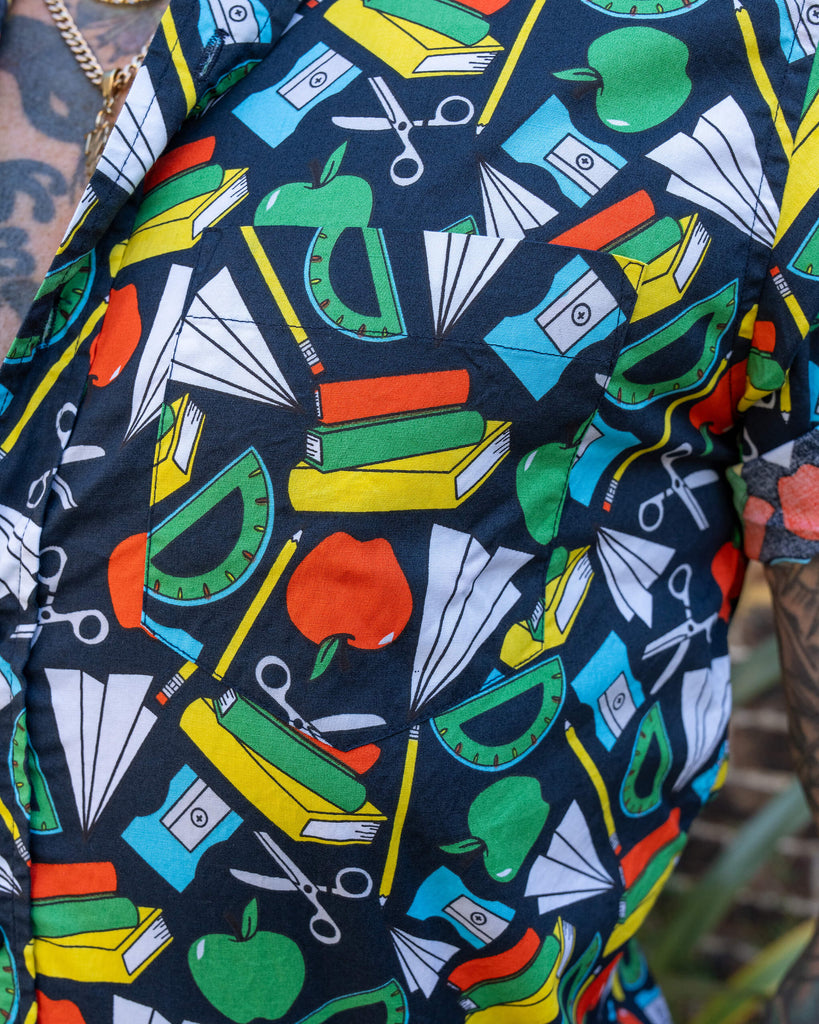 A close-up of a short-sleeve shirt with a school supplies pattern, worn by an older man with tattoos. The design includes books, apples, rulers, and scissors on a dark background.