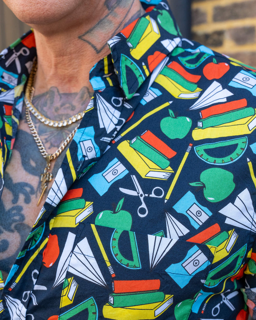 A close-up of a short-sleeve shirt with a school supplies pattern, worn by an older man with tattoos. The design includes books, apples, rulers, and scissors on a dark background.