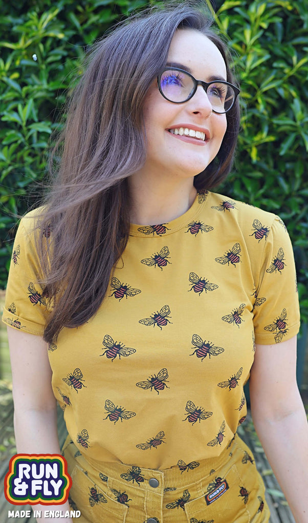 Amy is smiling and looking up into the distance whilst wearing black glasses and the Buzzy Bee Mustard Gold Tee.