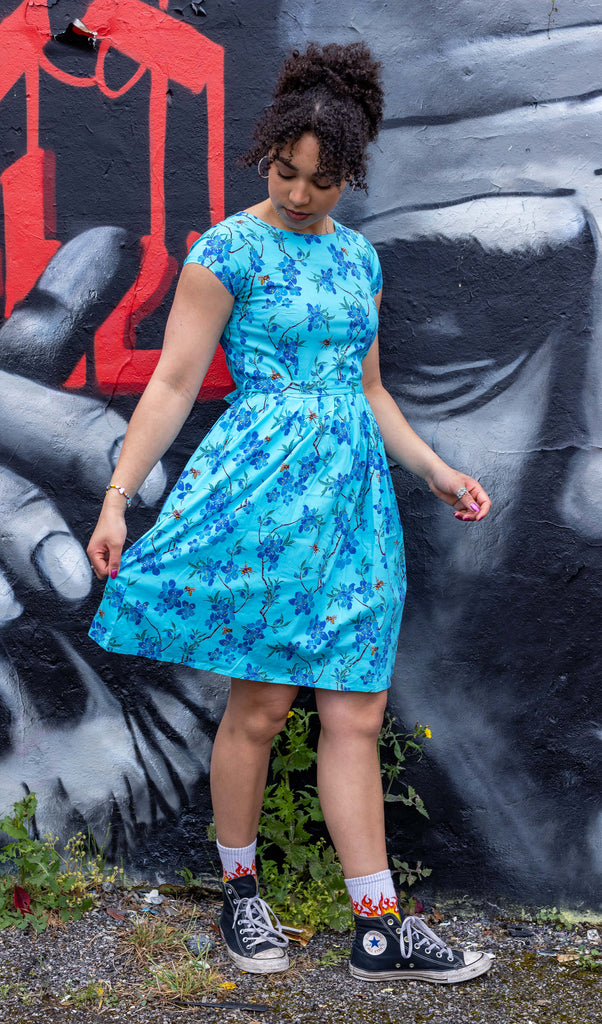 Georgia is stood in front of a graffiti wall wearing the cherry blossom black belted stretch tea dress with pockets with flame socks and black converse. She is facing forward posing