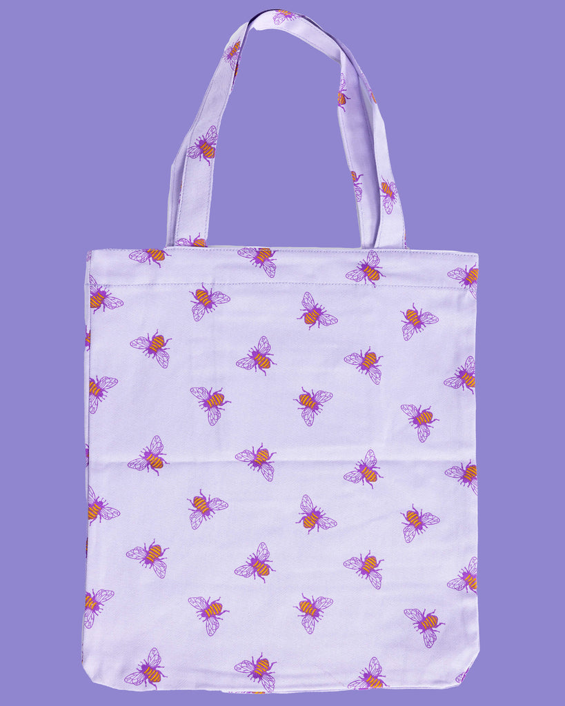 flat lay of a lilac tote bag with an all over print of yellow bumble bees with purple wings on a purple backdrop. There is a front pocket on the tote bag with Run&Fly logo on the front.