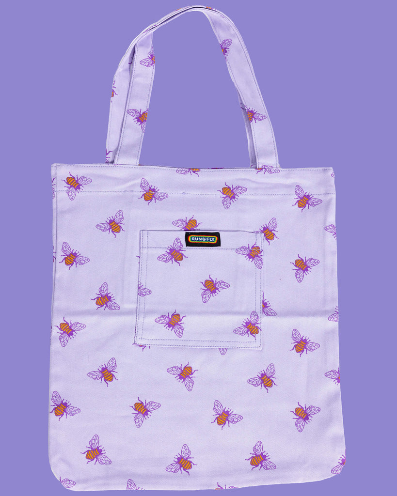 flat lay of a lilac tote bag with an all over print of yellow bumble bees with purple wings on a purple backdrop. There is a front pocket on the tote bag with Run&Fly rainbow logo on the front.
