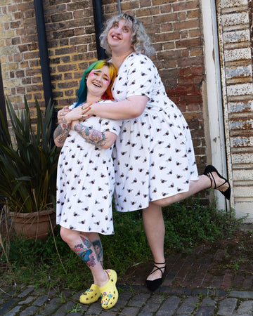 Two individuals standing and smiling in front of a brick wall, both wearing white oversized smock dresses with an all over bee print from slow, ethical fashion brand Run & Fly. One has rainbow-coloured hair and yellow Crocs, while the other has curly blonde hair and black shoes. They both have their hands in the dress pockets.